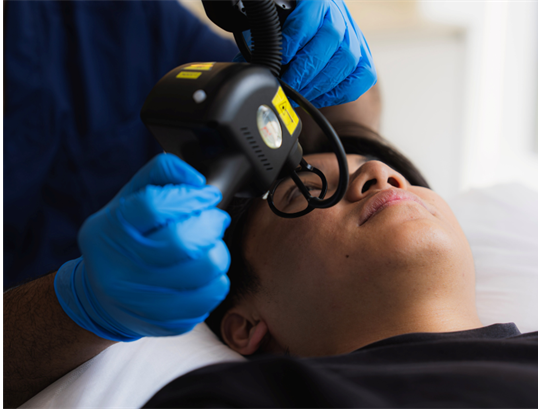 Clinician wearing gloves holding a handheld treatment device above the forehead of a man lying on a treatment couch.